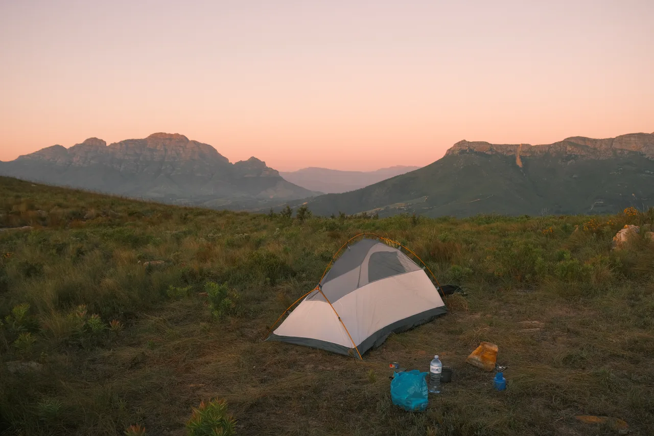 A tent pitched in mountain grass beneath a pink evening sky.