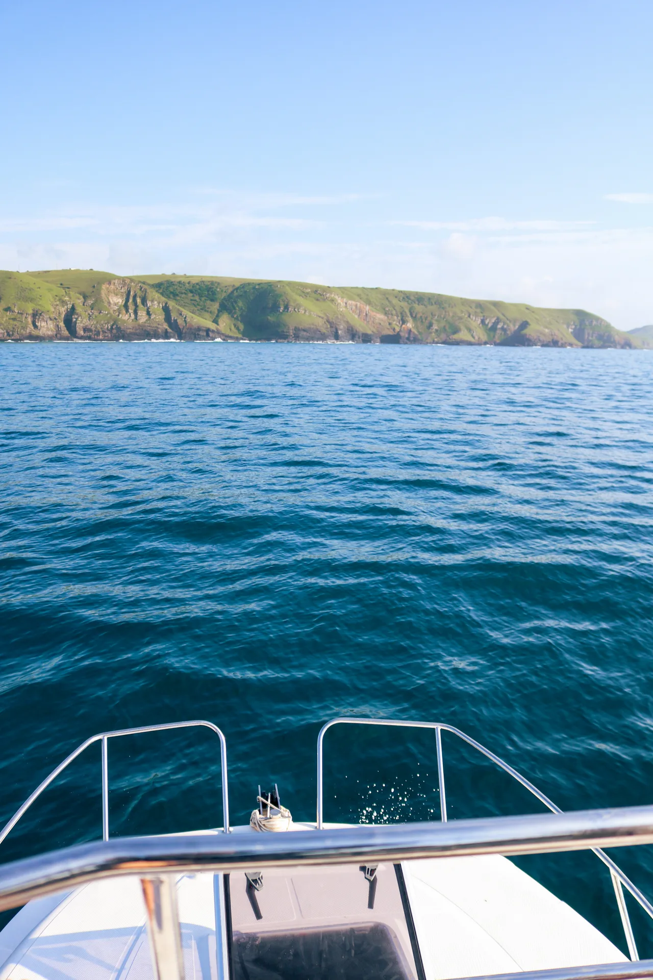 The bow of a boat crossing blue water toward distant cliffs.