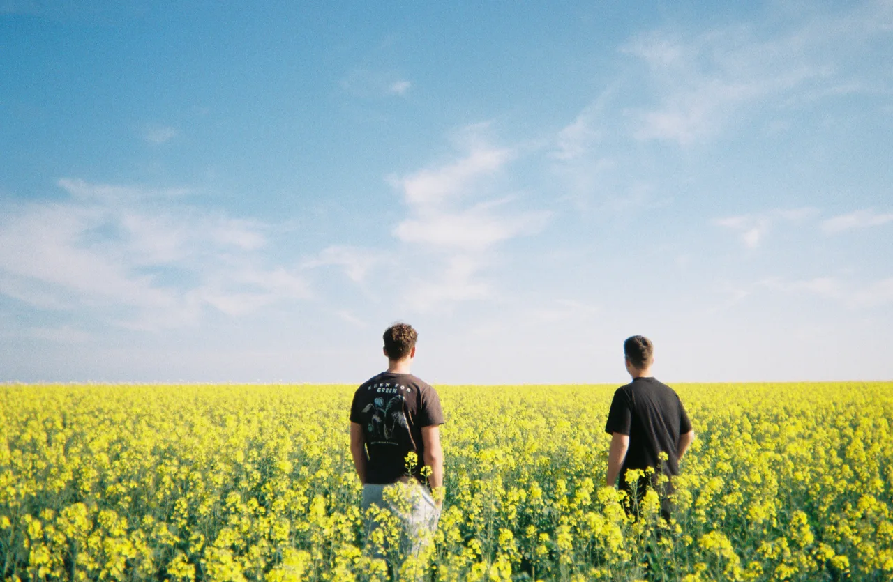 Two people standing in a field of yellow flowers beneath a blue sky.